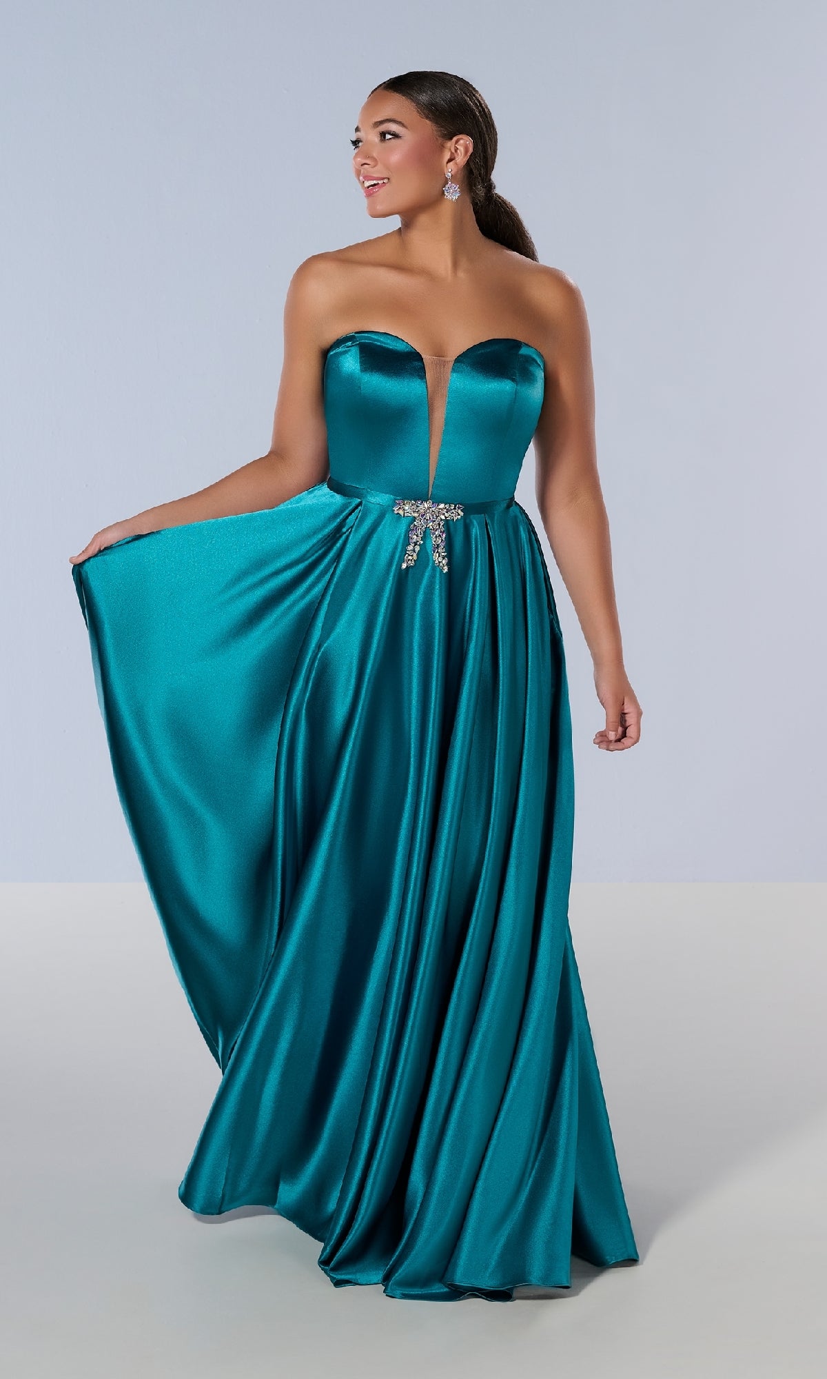A woman models the Tiffany 16219 long plus-size teal satin prom dress, featuring a strapless deep V neckline, jeweled waist accent, and a full floor-length skirt against a plain light background.